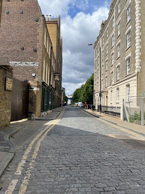 A narrow cobbled street in Hoxton, lined with old brick and stone buildings on either side. On the left, there is a dark wooden fence and a small gate, alongside a brick wall with visible signs of weathering. On the right, a large cream-colored building with multiple windows features black wrought iron railings along its base. The street is slightly curved and leads towards a vanishing point with trees and parked cars visible in the distance. Overhead, the sky is partly cloudy, casting diffused natural light across the scene. This setting exemplifies the challenging urban environment that Man with Van Hoxton often navigates during house removals, where careful planning is necessary to transport furniture and packed boxes through confined, historic streets without causing damage or disruption.