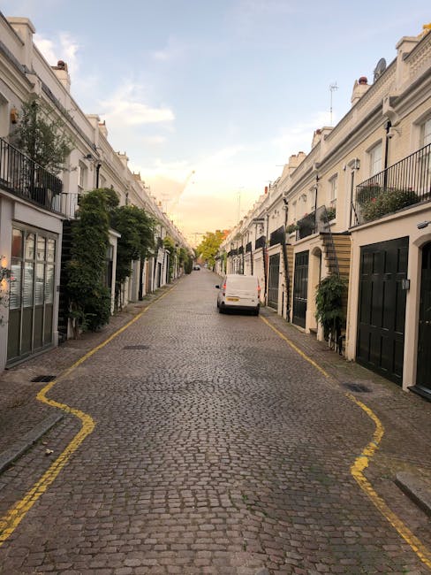 A narrow cobbled street in Hoxton, lined with old brick and stone buildings on either side. On the left, there is a dark wooden fence and a small gate, alongside a brick wall with visible signs of weathering. On the right, a large cream-colored building with multiple windows features black wrought iron railings along its base. The street is slightly curved and leads towards a vanishing point with trees and parked cars visible in the distance. Overhead, the sky is partly cloudy, casting diffused natural light across the scene. This setting exemplifies the challenging urban environment that Man with Van Hoxton often navigates during house removals, where careful planning is necessary to transport furniture and packed boxes through confined, historic streets without causing damage or disruption.