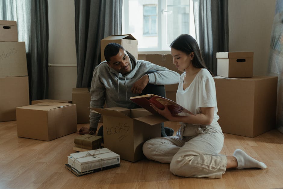 A young woman and a young man are inside a room with hardwood floors, surrounded by cardboard moving boxes, some labeled with words like 'BOOKS,' and wrapped packages. The woman is sitting on the floor wearing light-colored casual clothing and white socks, holding a red book. The man, dressed in a grey hoodie, is kneeling beside her and leaning over to look at the book. Behind them, there is a window with partially open curtains allowing natural light into the room. The boxes vary in size, with some stacked on top of each other, and appear to be prepared for a home relocation or furniture transport process managed by a professional removals service such as Man with Van Hoxton. The scene depicts a packing and moving activity, with the two individuals engaged in sorting or reviewing belongings as part of a household move.