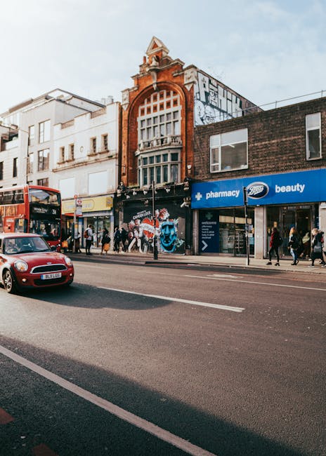 A street scene in Hoxton featuring a row of mixed-use buildings with retail shops on the ground floor and upper floors with windows. One building has a distinctive red brick facade with an ornate arched window at the top. On the ground floor, there are shops including a pharmacy and a beauty store, with signage clearly visible. Pedestrians are walking along the pavement, some entering or exiting the shops. A red double-decker bus and a small red car are moving along the street, which has designated lanes and parking. The scene is captured during daytime with natural lighting. This image illustrates the typical urban environment where house removals and furniture transport may involve navigating busy commercial areas, demonstrating the importance of planning and loading procedures carried out by professional moving services like Man with Van Hoxton for house relocations and local removals.