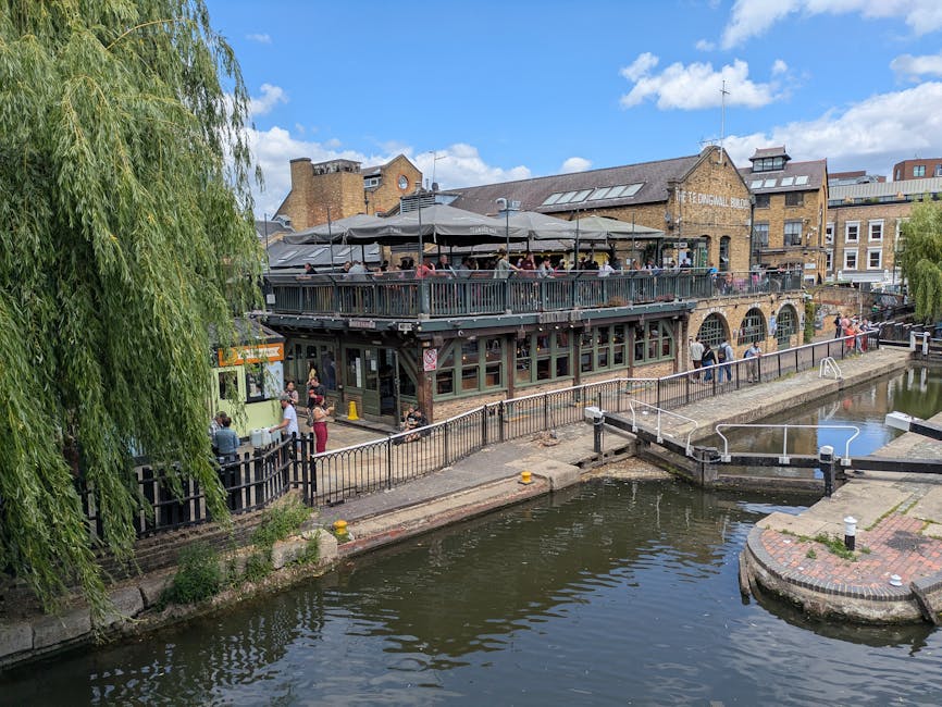 A view of a two-storey building situated alongside a canal, featuring a large rooftop terrace with outdoor seating under large umbrellas, populated with people. The building appears to be a restaurant or pub, with brick and glass construction, and has an arched ground-floor window design. Adjacent to the building, a narrow paved walkway runs along the canal, with a black metal fence separating pedestrians from the water. On the pavement, several people are waiting or walking, and some individuals are engaged in lifting or carrying boxes or furniture as part of a home relocation process. In front of the building, a small, vintage-style green delivery vehicle is parked, with a few items stacked nearby. The scene is illuminated by daylight, with some trees, blue sky, and scattered clouds visible, indicating a pleasant day for moving or delivery activities. Man with Van Hoxton's team is involved in the logistics of relocating household items during a furniture transport or packing and moving operation in Shoreditch's Regent's Canal area, close to the historic pub.