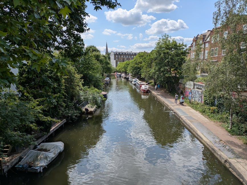 A canal scene in Shoreditch with calm water reflecting a partly cloudy sky, flanked on both sides by a paved promenade and a line of trees with lush green foliage. On the right, several parked vehicles are close to residential buildings, some of which feature graffiti on their walls, while pedestrians stroll along the path. On the left, there are boats moored along the canal bank, including one covered with a tarp. The background shows a church spire rising above the trees and buildings. This image relates to house removals and relocating furniture in urban settings, illustrating the environment where transportation and packing services, such as those provided by Man with Van Hoxton, are often needed for home relocation or furniture transport.