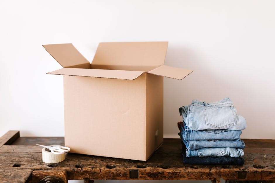 A plain cardboard box with its flaps open is positioned on a rustic wooden surface in a well-lit room, accompanied by a neatly folded stack of jeans in varying shades of blue beside it. A roll of packing tape with a tape dispenser is also placed nearby on the wood, indicating ongoing packing or preparation for a house move. The background consists of a plain, light-colored wall, and the scene captures the initial stages of home relocation, with furniture and packing materials suggesting a furniture transport or packing and moving process. The presence of the box, packaging tape, and folded clothing reflects typical elements involved in house removals carried out by [COMPANY_NAME]. This image emphasizes the logistical aspects of furniture transport and packing services in a home relocation context, consistent with practices discussed in the page titled 'Estate moves on Kingsland Road: delivery and access tips, HOXTON' at [PAGE_URL].