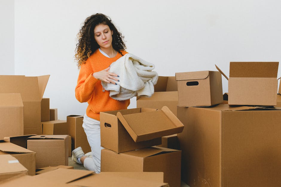 A young woman with curly hair, dressed in a bright orange sweater and white pants, is kneeling on the floor amidst a large number of unpacked cardboard boxes of various sizes, some open and some sealed with packing tape. She is holding a soft, cream-colored blanket or cloth while examining it, surrounded by a clutter of moving materials inside a plain, well-lit room with white walls. The boxes are arranged around her, indicating an active home relocation or packing process typical of furniture transport and moving logistics. Some boxes feature cut-out handles, and others are stacked or opened, with visible packing items such as bubble wrap and paper padding. The environment suggests an ongoing packing process before or during a house move, and the presence of these boxes and materials highlights the detailed organization involved in household packing and moving activities, complemented by the focus on careful handling and preparation for a furniture transport or home relocation service, as offered by Man with Van Hoxton.