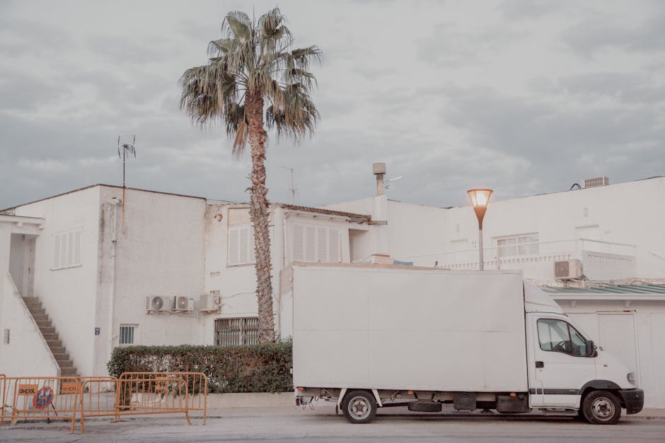 A plain cardboard box with its flaps open is positioned on a rustic wooden surface in a well-lit room, accompanied by a neatly folded stack of jeans in varying shades of blue beside it. A roll of packing tape with a tape dispenser is also placed nearby on the wood, indicating ongoing packing or preparation for a house move. The background consists of a plain, light-colored wall, and the scene captures the initial stages of home relocation, with furniture and packing materials suggesting a furniture transport or packing and moving process. The presence of the box, packaging tape, and folded clothing reflects typical elements involved in house removals carried out by [COMPANY_NAME]. This image emphasizes the logistical aspects of furniture transport and packing services in a home relocation context, consistent with practices discussed in the page titled 'Estate moves on Kingsland Road: delivery and access tips, HOXTON' at [PAGE_URL].