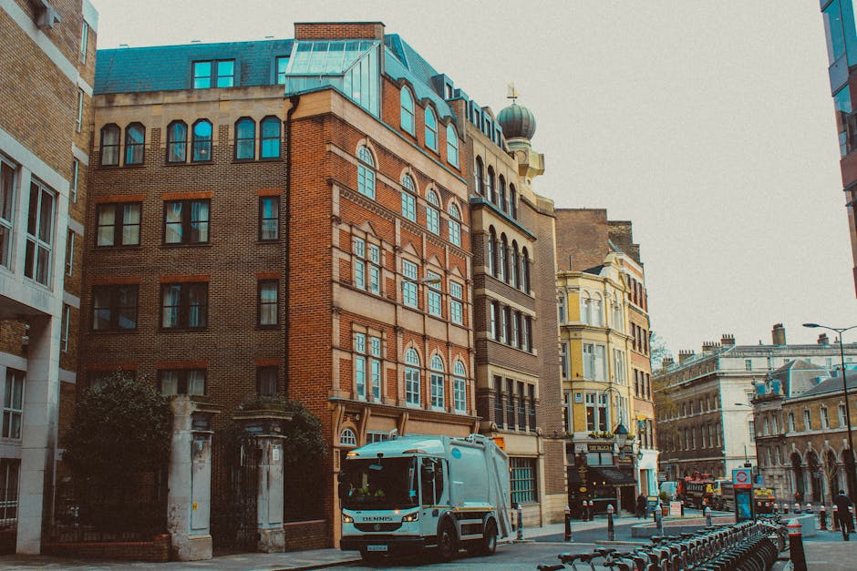 A white courier van from Man with Van Hoxton is parked on a busy city street in Hoxton, surrounded by multi-storey buildings featuring a mix of brick and Victorian architectural styles, with large windows and decorative facades. The van is positioned close to a building entrance, with cardboard boxes and packing materials visible nearby on the pavement, indicating a home relocation or furniture transport process. The scene is well-lit with natural daylight, and adjacent structures include modern and historic designs, reflecting an urban environment where residential move logistics and loading activities take place, often influenced by local parking restrictions like Hackney CPZ. The van is part of the ongoing loading or unloading operation, with some boxes possibly being carried or prepared for delivery as part of the moving services offered by [COMPANY_NAME], supporting house removals and packing and moving tasks in the area.