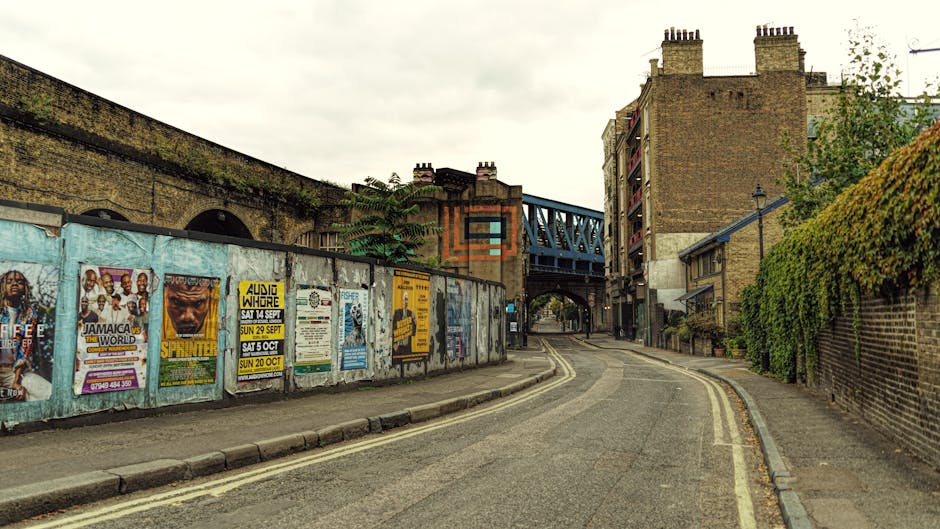 A narrow, curved street scene showing a pavement on the left side with a low brick wall covered in colorful posters and advertisements, including event promotions and music posters. Behind the wall, t