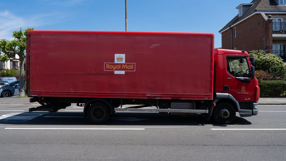 A large red Royal Mail delivery van parked on the side of a residential street during daytime, with a building featuring balconies and greenery visible in the background. The van's cargo area is closed, with no visible objects inside. The vehicle is positioned in a designated parking lane, with white lines marking the space, and the pavement is adjacent to the street. The side featuring the Royal Mail logo displays a white box with a gold crown and red lettering. The cab of the van has a black front bumper and black side mirrors, and the vehicle appears to be a medium-sized commercial delivery truck. The environment is well-lit with clear weather, suggesting regular home delivery or courier operations, relevant to moving or packing services supported by Man with Van Hoxton.