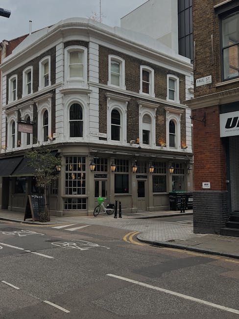 The image depicts a three-storey corner building on Shoreditch High Street, with a white facade and large windows, some of which are open. The ground floor features a black-framed entrance with multiple panes of glass, and outdoor lighting fixtures above the windows. In front of the building, there is a bicycle parked against a black bollard, and a green flag attached to the bicycle. The street shows marked bicycle lanes and double yellow lines along the curb. Adjacent to the building, part of a modern structure with glass and brick is visible, along with a black brick wall and a street sign indicating 'Paul St'. The setting appears to be during daylight hours with natural light illuminating the scene. This environment is typical for home relocation and furniture transport activities, with the street suitable for loading and unloading by [COMPANY_NAME], as part of its removals and moving services on Shoreditch High Street.