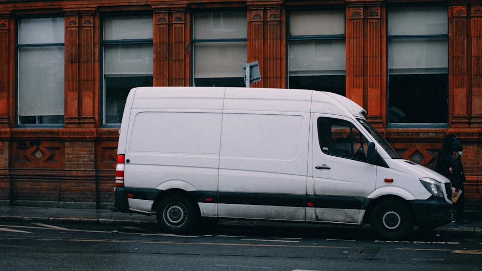 A white delivery van parked on a street in front of a red-brick building with decorative stone window frames and intricate masonry details. The van's side profile shows it is a medium-sized commercial vehicle, with black bumper and side mirror, and black wheel arches. The street has a double yellow line along the curb, indicating parking restrictions, and a pedestrian is walking past the van on the sidewalk. The building behind features three large windows, partly covered by blinds, and the entrance is not visible. The environment suggests an urban area suitable for home relocation or furniture transport services, which Man with Van Hoxton provides. The scene captures the loading process of moving furniture or boxes, with the van positioned close to the building for efficient loading or unloading, and a trolley or box might be nearby for moving items. The lighting is natural, likely during daytime, emphasizing the practical aspect of house removals, packing, and furniture transport in a city setting.