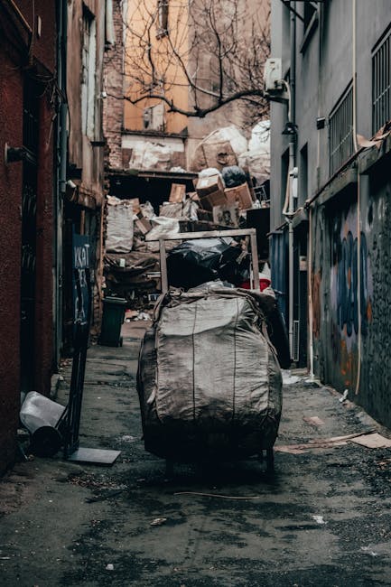 A large, grey, fabric sack filled with bulky waste material is positioned on a wheeled trolley in a narrow urban alleyway between two buildings. The alley is cluttered with various discarded items including cardboard boxes, plastic bags, and construction debris piled against a fence at the end of the alley. The ground is paved with dark asphalt and shows signs of dirt and wear. To the right, the side of a grey building features graffiti artwork, while on the left, a brick wall displays garage doors and windows. Overhead, leafless tree branches extend across the scene, with soft natural light illuminating the area. The image captures a typical home clearance process involving collection and transport of waste, associated with removals services provided by Man with Van Hoxton, and highlights the logistical aspects of furniture transport and loading during a house move or clearance operation.