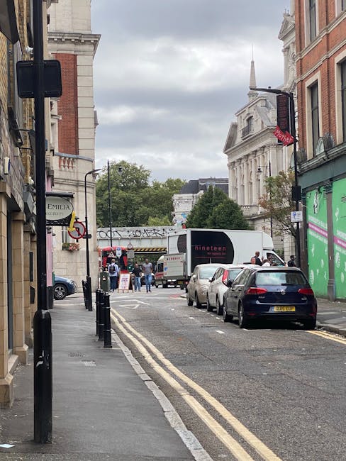 A narrow urban street scene in Hoxton, showing a row of parked cars along the right side and black bollards lining the pavement on the left. In the background, a white moving truck with the logo 'dinetea' is being loaded or unloaded, with several people visible around it. The street is bordered by old brick and stone buildings, some with shopfronts and signage, including a green storefront and a red awning. Overhead, there is a bridge or gantry with graffiti, and a historic white building with classical architecture is visible beyond the trees. The sky is overcast, creating a diffused light over the scene, which exemplifies the typical urban environment where a house removal or furniture transport could take place as part of a home relocation process, with visible packing materials and moving activities involved, orchestrated by Man with Van Hoxton.