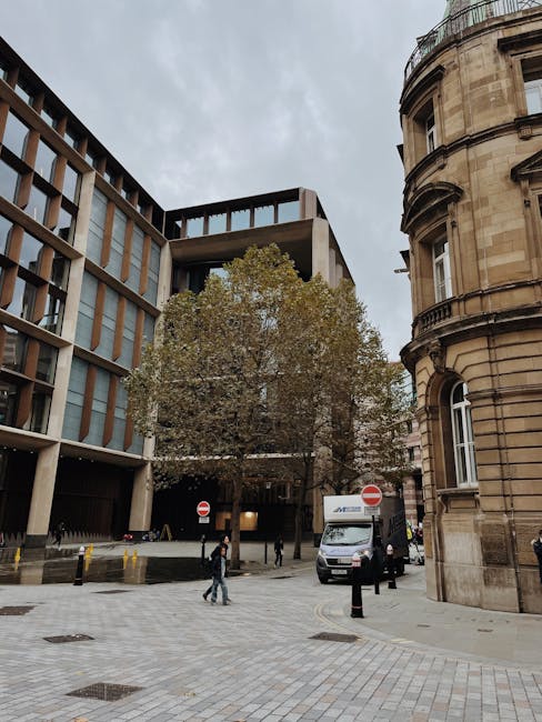A street scene in Hoxton with a mix of modern and historic architecture, including a multi-storey office building with a concrete facade and large windows on the left, and a rounded, terraced stone building on the right. In the foreground, there is a paved cobblestone area with black bollards, and a large deciduous tree with brown leaves, indicating autumn. Several pedestrians are walking across the street, some near a white delivery van parked close to the curb, which is used for home relocation or furniture transport. The van is positioned near a building entrance, and the scene is illuminated by natural light on a cloudy day, reflecting typical urban moving logistics context, as seen in professional removals services like those provided by Man with Van Hoxton.