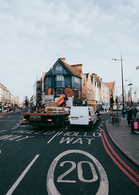 A narrow urban street scene in Hoxton, showing a row of parked cars along the right side and black bollards lining the pavement on the left. In the background, a white moving truck with the logo 'dinetea' is being loaded or unloaded, with several people visible around it. The street is bordered by old brick and stone buildings, some with shopfronts and signage, including a green storefront and a red awning. Overhead, there is a bridge or gantry with graffiti, and a historic white building with classical architecture is visible beyond the trees. The sky is overcast, creating a diffused light over the scene, which exemplifies the typical urban environment where a house removal or furniture transport could take place as part of a home relocation process, with visible packing materials and moving activities involved, orchestrated by Man with Van Hoxton.