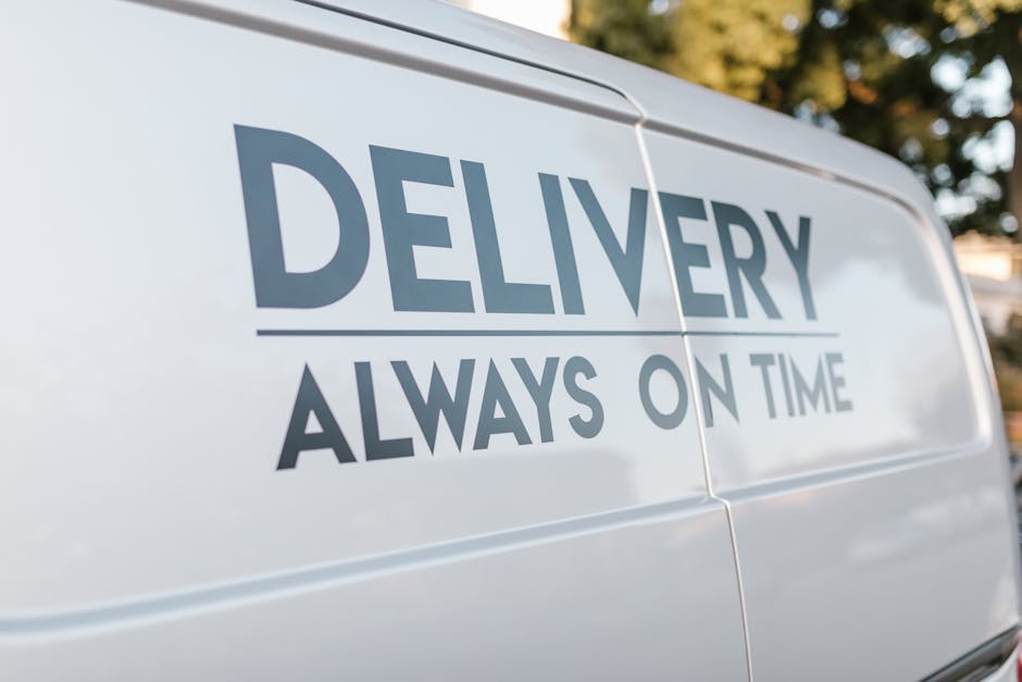 A white moving van with the logo 'MOVING COMPANY LOCAL & LONG DISTANCE' parked in front of a modern, two-story residential building during daylight. Inside the open driver's door, a person in casual c