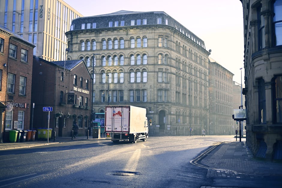 A white box truck with the logo of Man with Van Hoxton is parked on a street near a multi-storey historic building with arched windows. The truck is positioned at the edge of the pavement, close to th